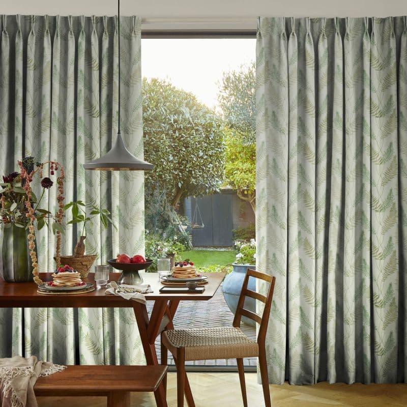 Pleated floor-length curtains with pale green fern print, drawn aside to reveal a glass door, filtering daylight into a dining nook with a wooden breakfast table and garden view.