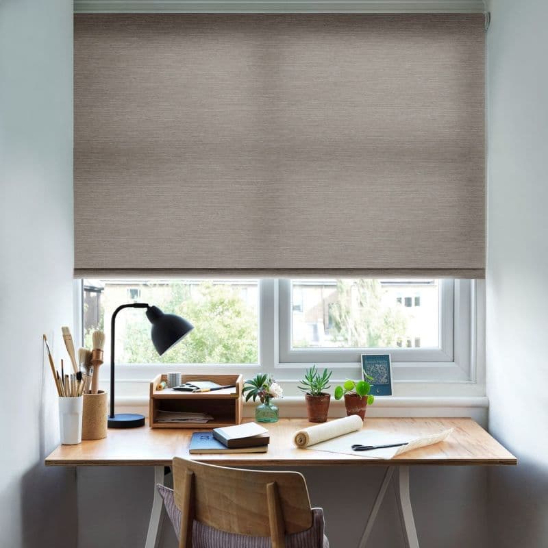 Beige textured roller blind lowered nearly fully, filtering daylight; a wooden desk beneath holds a black desk lamp, art brushes, notebooks, small potted plants, and a suburban window view.