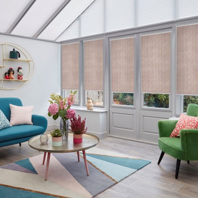 Beige textured roller blinds (lowered over tall patio windows and doors) and white pleated skylight blinds partially diffuse daylight in a bright conservatory living room with colorful sofas.
