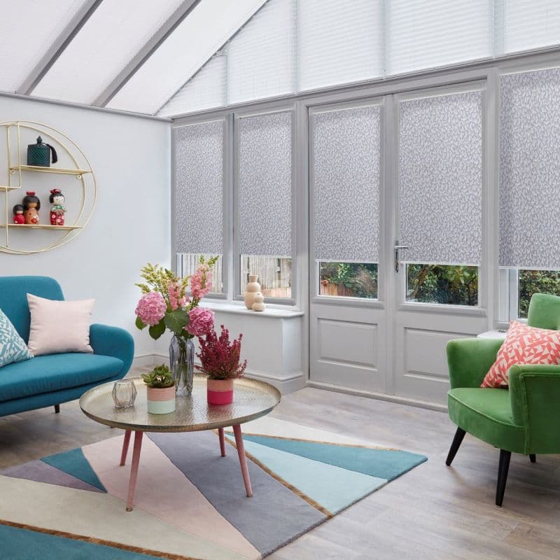 Patterned grey roller blinds cover tall conservatory windows and door, partially lowered and diffusing daylight; sunroom with pastel sofa and armchair, round coffee table, floral arrangements, geometric rug.
