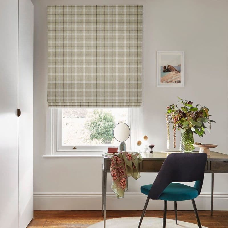 Beige plaid Roman blind over a sash window, fully lowered and softly filtering daylight; a wooden desk with teal chair and vase of flowers beneath, neutral-walled room, garden view.