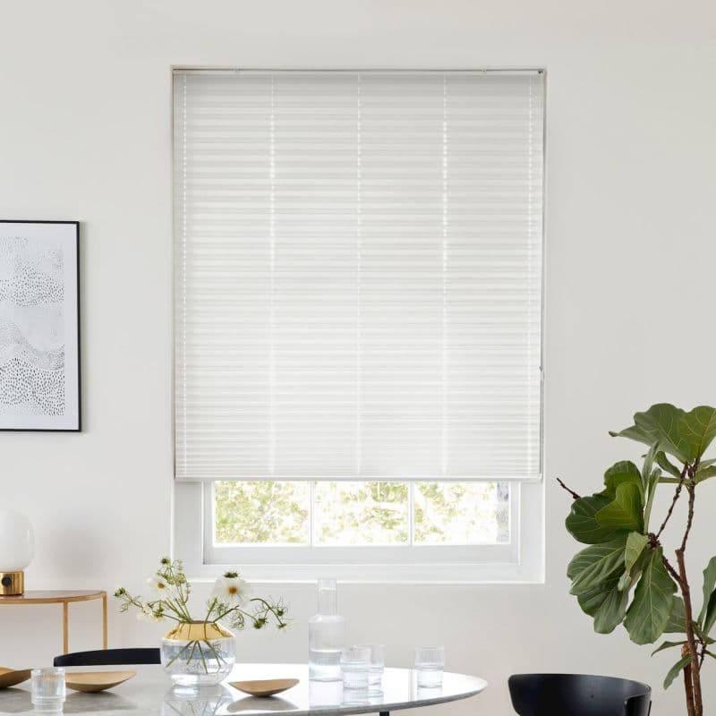 White pleated blind, fully lowered and translucent, filtering daylight over a recessed window in a bright dining nook with table, glass vase of flowers, framed art and a fiddle‑leaf fig.