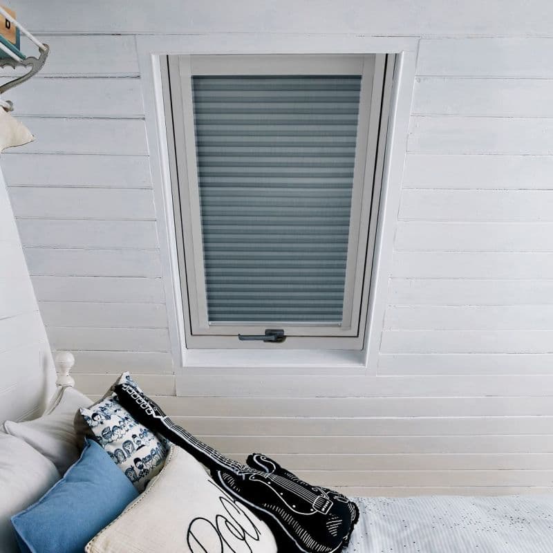 Pleated skylight blind, fully lowered in a centered white-framed roof window with blue-grey horizontal stripes; filtering soft daylight in a white-paneled attic bedroom with bed, pillows and guitar-shaped cushion.