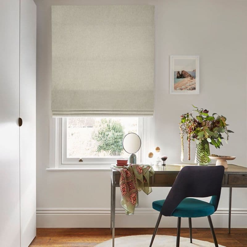 Beige fabric Roman blind lowered over a sash window, softly filtering daylight; in front, a wooden desk with teal-upholstered chair, small mirror, vase of foliage, and neutral-painted room.