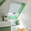 White pleated cellular skylight blind, partially lowered inside the roof window, filtering soft daylight into an attic bathroom with green trim, white herringbone tiles, sink, bathtub, plant, and bath tray.