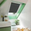 Dark gray pleated skylight blind (on an angled roof window), partially lowered and filtering daylight; bathroom with green-painted window trim, white herringbone tiles, sink, bathtub, potted plant, and toiletries.