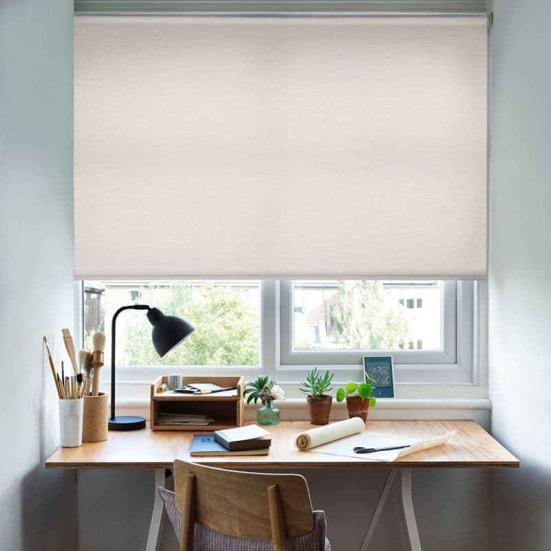 A pale beige fabric roller blind, fully lowered, diffusing daylight through the window. On the sill and desk: a wooden workspace with lamp, brushes, small potted plants, notebooks, chair.