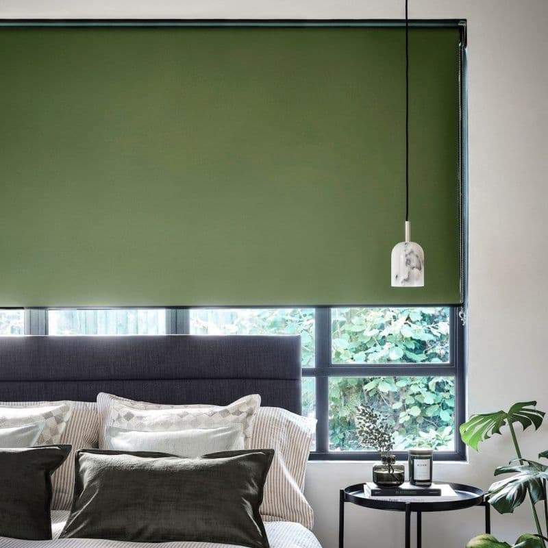 Green roller blind lowered across a window, its fabric blocking light while a narrow bottom gap reveals leafy garden; bedroom setting with upholstered headboard, pillows, bedside table, hanging pendant lamp.