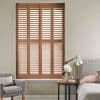 Wooden four-panel plantation shutters with horizontal louvers, closed and slightly angled to filter daylight, covering a tall window in a neutral bedroom corner—bed, bedside table, lamp and armchair.