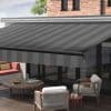 Striped retractable awning (dark gray, diagonal stripes, drop valance) extended over a patio, shading glass doors and outdoor seating on a wooden deck in daylight.