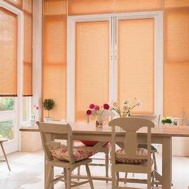 Horizontal slatted peach blinds covering tall central and side windows, closed and filtering warm daylight; dining table with chairs, floral cushions and vases of flowers in a cozy breakfast nook.