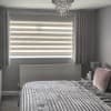Zebra roller blind with alternating opaque and sheer horizontal bands lowered across a wide window, flanked by pleated grey curtains at the sides; filtering soft daylight into a grey-toned bedroom.