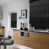 Black horizontal blinds cover a large window and smaller door window, fully lowered and closed, filtering light in a modern kitchen with light wood cabinets, black countertop and bar stools.