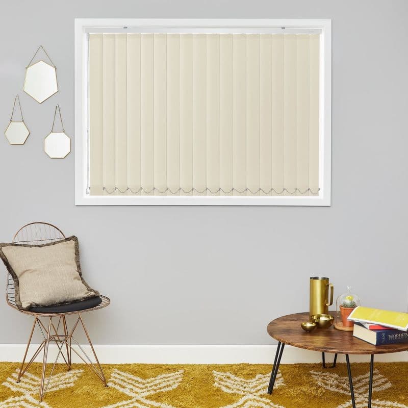 Beige vertical blinds, closed and hanging straight with chain-linked bottoms, softly diffusing daylight; set in a grey-walled living room with wire chair, mustard rug, wooden side table, books, gold cup.