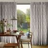 Pleated, pale-grey patterned curtains with feather motifs, pulled aside to reveal a central glass door; sunlight filters through onto a breakfast-set wooden dining table, pendant lamp, chair, and garden beyond.