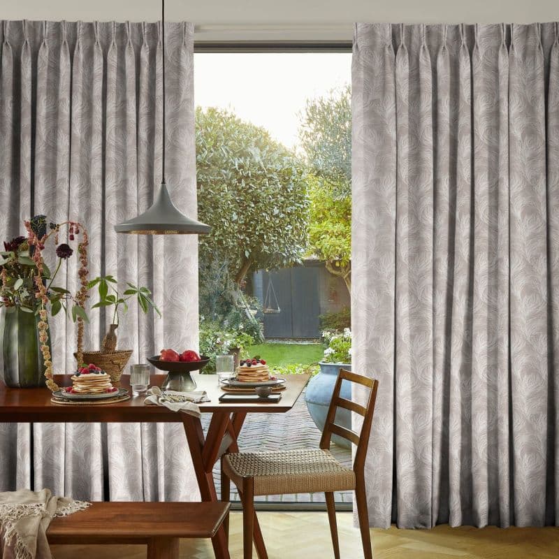 Pleated, pale-grey patterned curtains with feather motifs, pulled aside to reveal a central glass door; sunlight filters through onto a breakfast-set wooden dining table, pendant lamp, chair, and garden beyond.