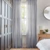 Sheer gray floor-length curtains hung either side of a sash window, filtering soft daylight and parted to show foliage outside; bedroom with dark paneled wardrobe and bedside table.