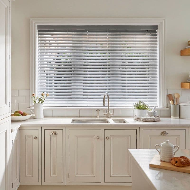 White horizontal Venetian blinds fully covering the window, slats slightly tilted, filtering soft daylight; above a stainless sink on a white marble countertop with cream cabinets, potted herb and utensils.