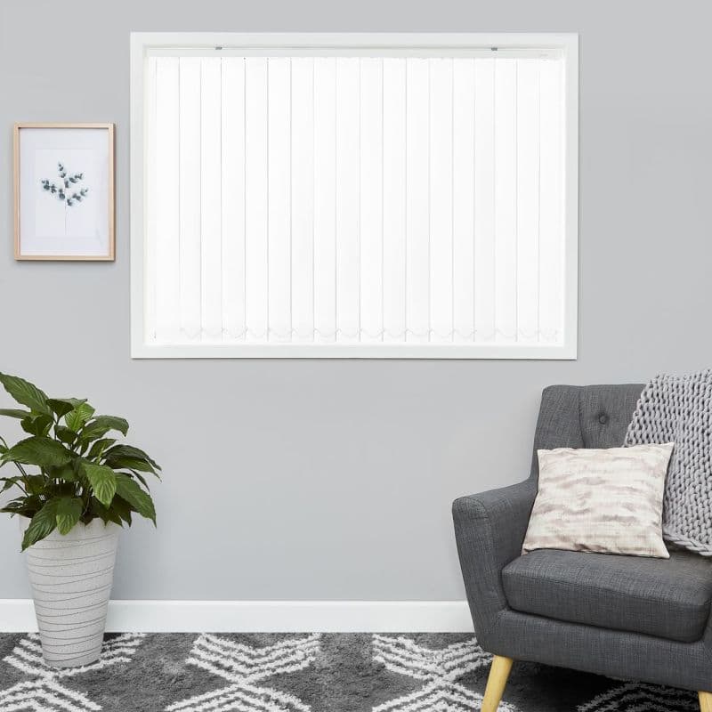 White vertical blinds, closed—evenly spaced slats with scalloped weighted bottoms—filtering bright daylight; mounted in a gray-walled living room above a patterned rug, beside a potted plant and gray armchair.