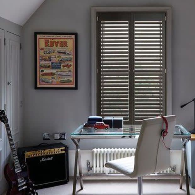 Wooden interior plantation shutters cover a double window, closed with slats angled to filter daylight; set above a glass desk in a grey home office with a chair, guitar and amplifier.
ROVER
Marshall