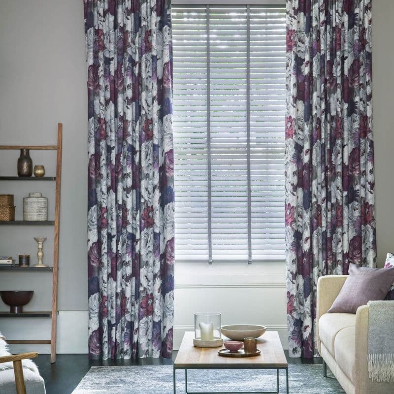 Floor-length pleated floral curtains (purple, white) frame a central window with closed horizontal blinds; curtains hang to the sides while blinds filter soft daylight into a cozy living room.