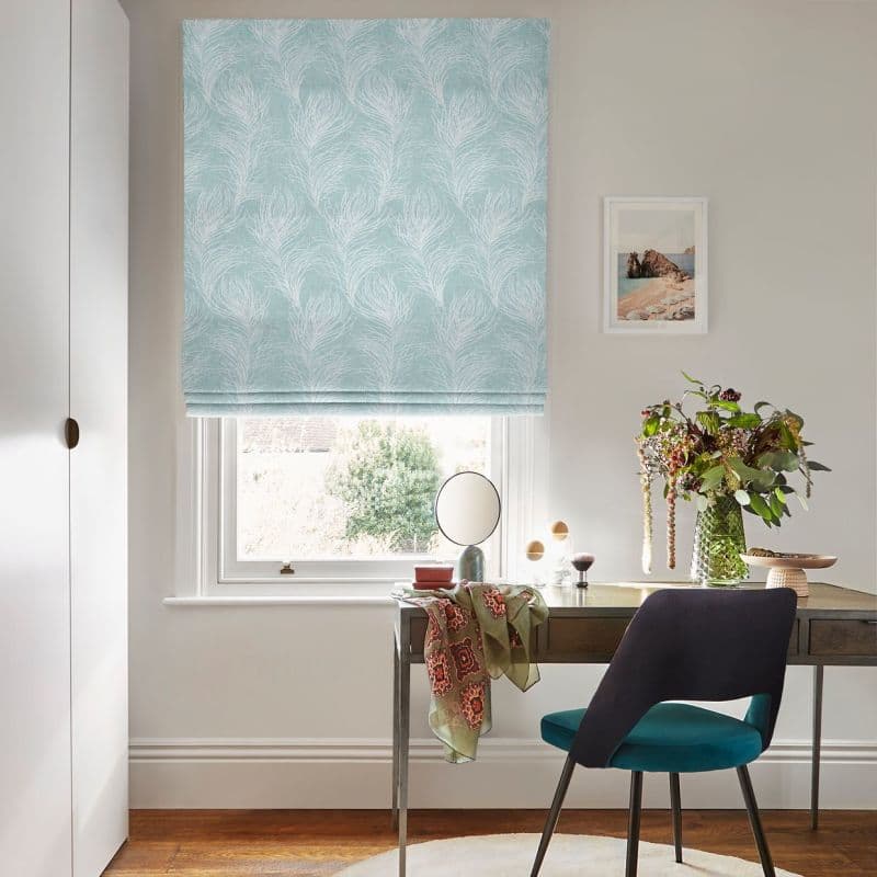 A pale teal Roman blind with white feather motif, lowered and softly folded, filters daylight over a small desk and teal chair in a bright, neutral home study with a vase of flowers.