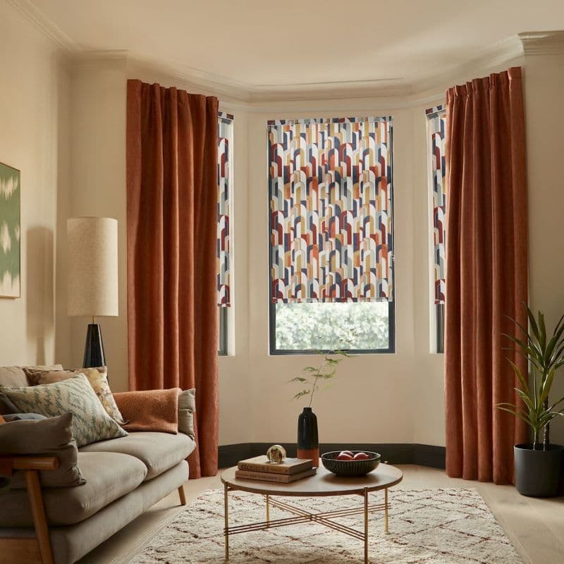 Patterned roller blind and rust velvet curtains in a bay window: blind lowered with geometric arches, curtains drawn to sides; warm living room with sofa, coffee table, rug.