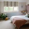 Cream Roman blind with irregular dark spots lowered across a three-pane window, partially filtering daylight in a pastel bedroom with bed, pink throw, potted plant and stuffed-animal hammock.