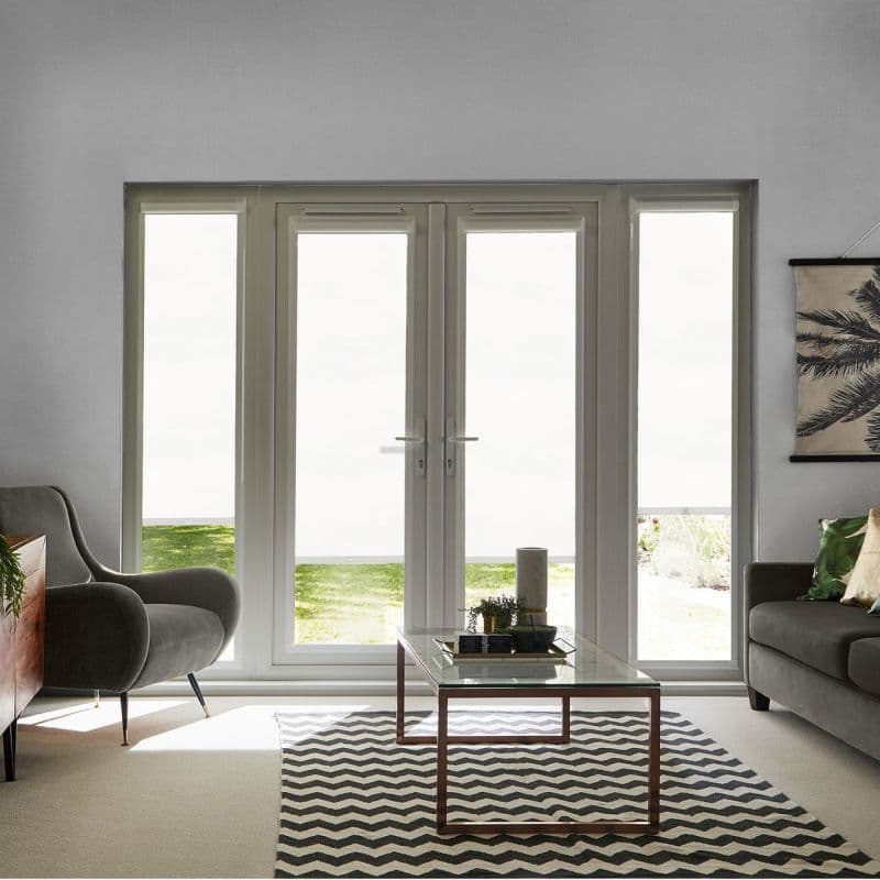 White translucent roller blinds covering tall French doors and sidelights, fully lowered, filtering bright daylight; living room with armchairs, glass coffee table on chevron rug and small garden glimpse below.