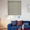 Beige narrow‑pleated blind, lowered to cover most of the window with a bottom gap, filtering daylight. Bright living room with blue sofa, cushions, floor lamp and framed art; rooftop beyond.