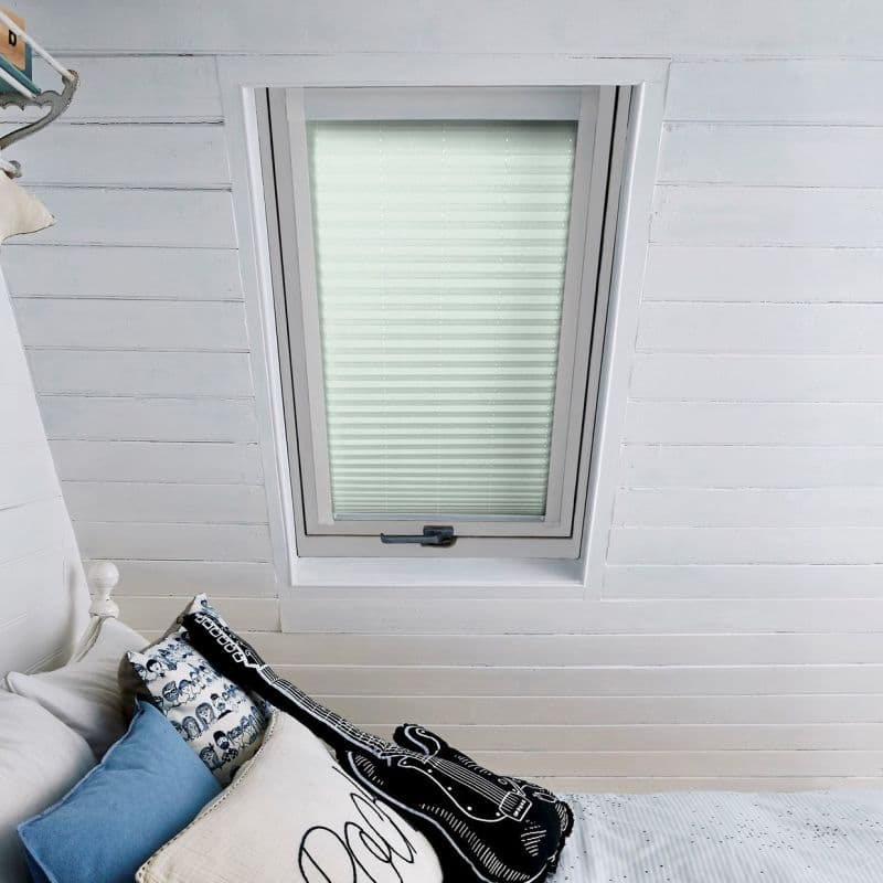 White pleated (cellular) blind fully lowered in a skylight window, gently diffusing light; set in a white-painted tongue-and-groove attic ceiling above a bed with pillows and a guitar-shaped cushion.