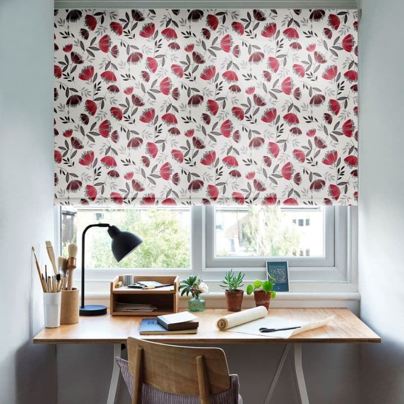 Patterned roller blind with white background and red-gray floral print, fully lowered and filtering daylight across a tidy wooden desk with lamp, plants, books, and a suburban window view.