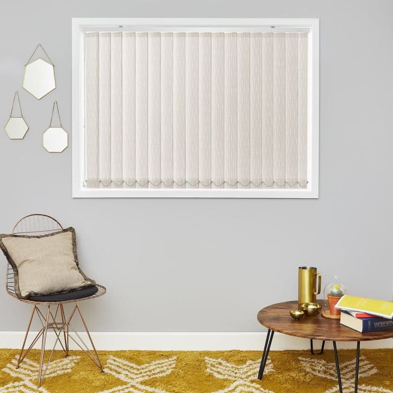 Beige vertical fabric blinds cover window, closed and softly filtering light in a white frame; grey living room wall with wire chair and cushion left, round wooden coffee table right.