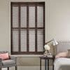 Dark wooden plantation shutters—four tall louvered panels covering the window—are closed with slats slightly tilted, filtering soft daylight into a neutral-toned bedroom with bed, armchair, and bedside table.