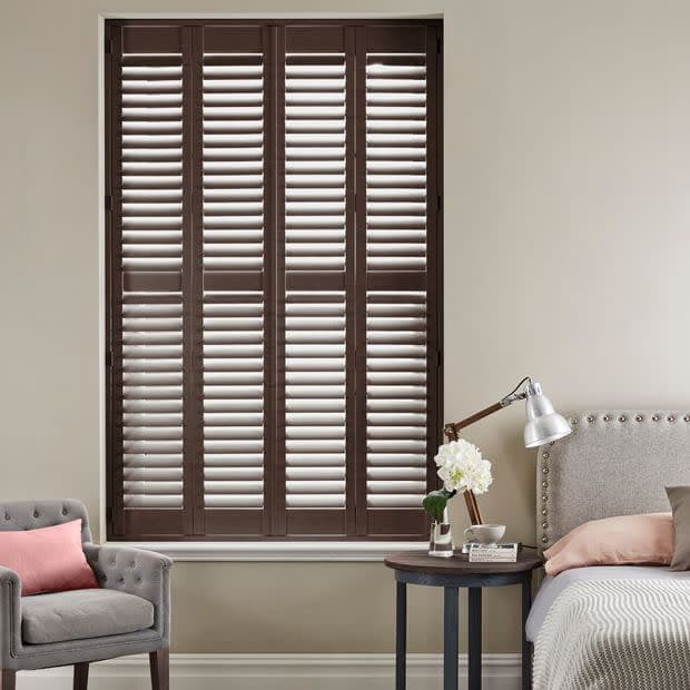 Dark wooden plantation shutters—four tall louvered panels covering the window—are closed with slats slightly tilted, filtering soft daylight into a neutral-toned bedroom with bed, armchair, and bedside table.
