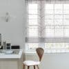 Sheer patterned Roman blind (leaf-chevron motif), lowered to mid-height and filtering soft daylight through a paneled window; minimal white home office with wooden chair, desk, pendant paper lamp, tree view.