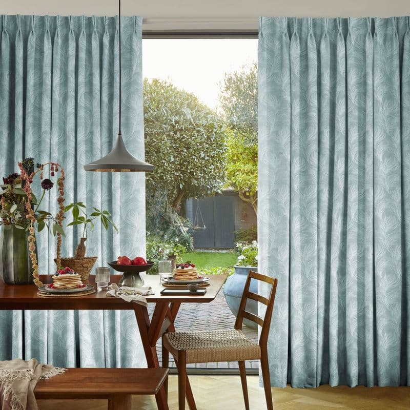 Floor-length pleated pale-blue patterned curtains pulled aside, framing a glass patio door and softly filtering daylight; a wooden dining table set for breakfast, pendant lamp, chairs, and sunny garden view.