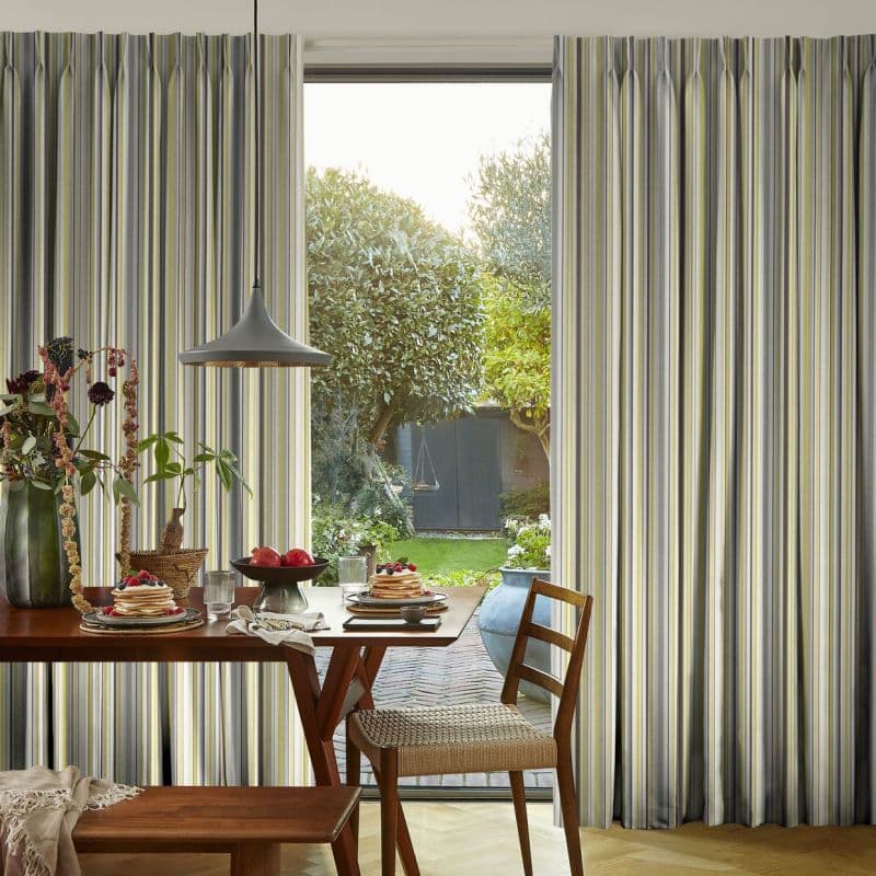 Floor-to-ceiling pleated striped curtains (gray, white, yellow) frame a central glass door, parted to reveal sunlight and a leafy backyard; foreground dining table set with pancakes, pendant lamp, wooden chairs.