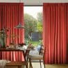 Red pleated floor-length curtains, slightly parted, framing a glass door and admitting daylight and a leafy garden; a wooden dining table with pancakes, pendant lamp and chairs in foreground.