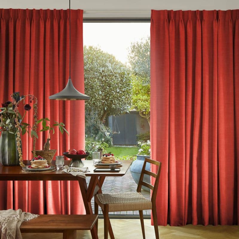 Red pleated floor-length curtains, slightly parted, framing a glass door and admitting daylight and a leafy garden; a wooden dining table with pancakes, pendant lamp and chairs in foreground.
