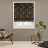 Patterned dark Roman shade with geometric diamond print, lowered over a window, partially filtering daylight; in a taupe living room with mid-century chair, cushion, small side table and potted plant.