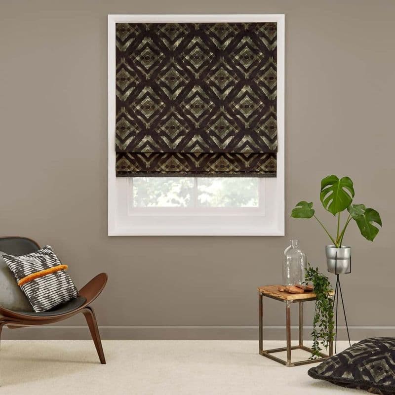 Patterned dark Roman shade with geometric diamond print, lowered over a window, partially filtering daylight; in a taupe living room with mid-century chair, cushion, small side table and potted plant.