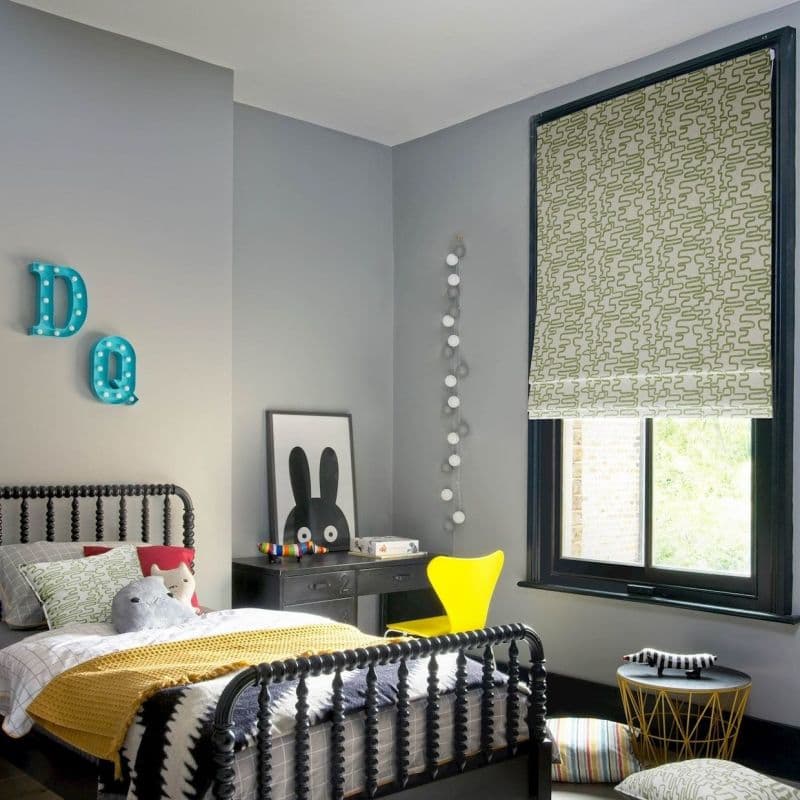 Green-and-white patterned Roman shade lowered over a black-framed window, filtering daylight; child’s grey-painted bedroom with black spindle bed, yellow chair, desk, plush toys and string lights.