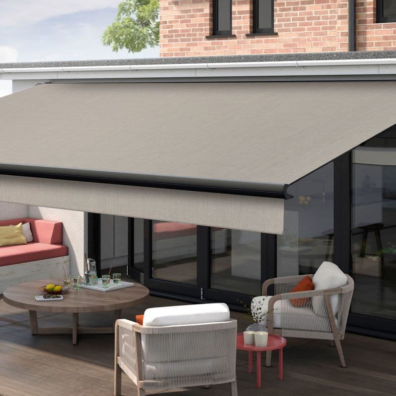 Beige retractable fabric awning with black front bar and hemmed valance, fully extended and shading glass doors and patio; modern outdoor seating, round coffee table, brick upper wall, daylight.
