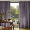 Pleated mauve floor-length curtains, drawn aside to reveal a central opening; framing and filtering soft daylight in a dining area with a wooden table and leafy garden view.