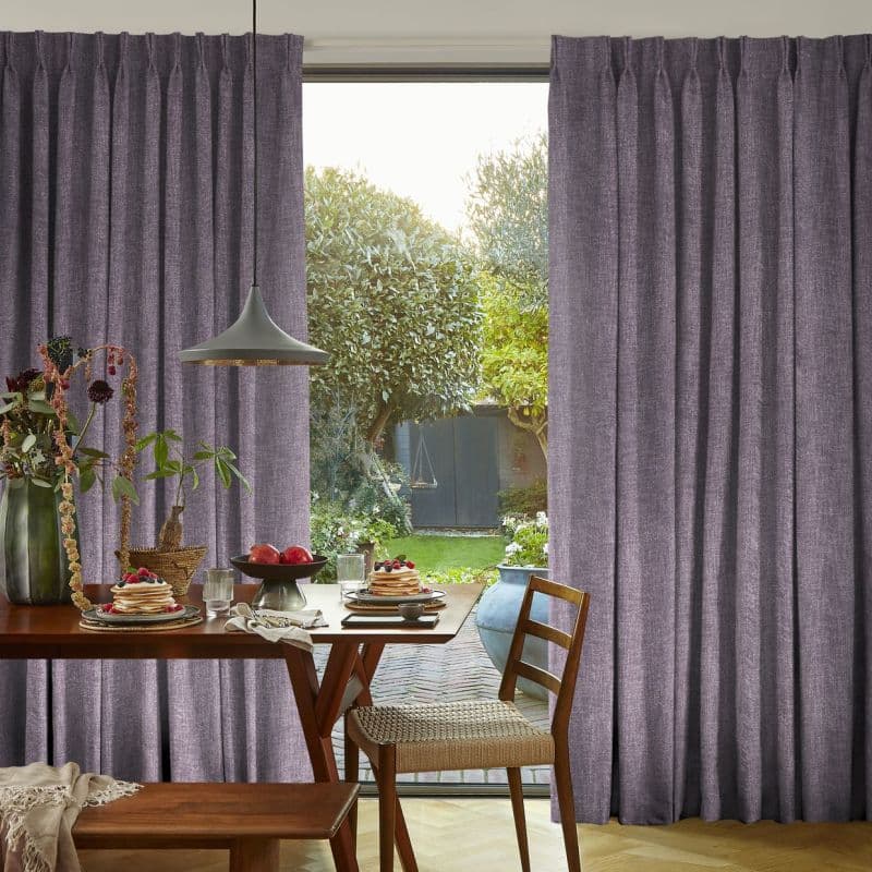 Pleated mauve floor-length curtains, drawn aside to reveal a central opening; framing and filtering soft daylight in a dining area with a wooden table and leafy garden view.