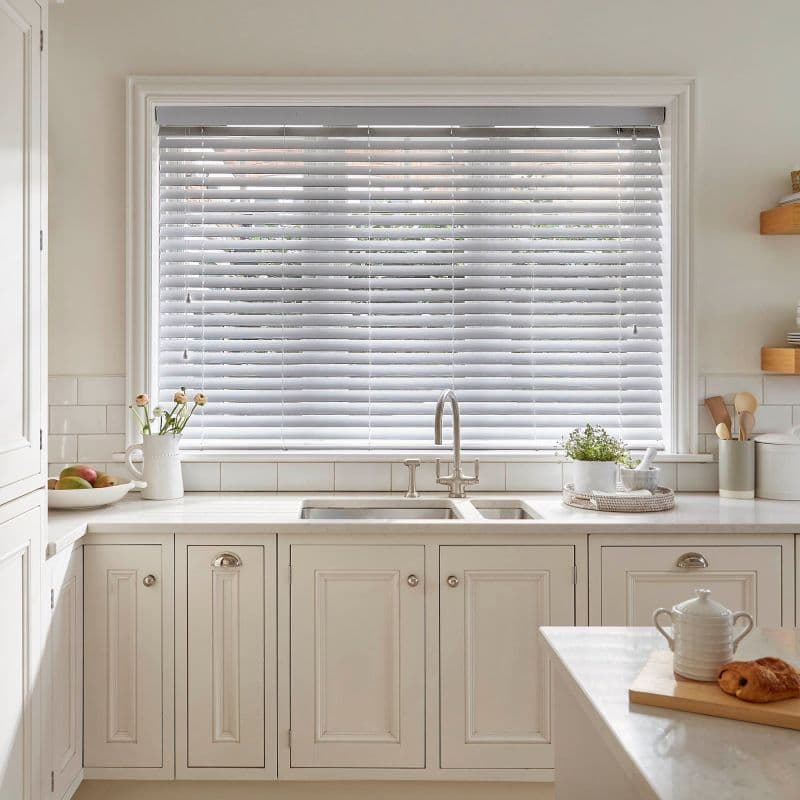 White horizontal Venetian blinds across a large kitchen window, slats partially closed and filtering soft daylight; stainless sink and cream cabinetry beneath, countertop with plants, utensils, and breakfast items.