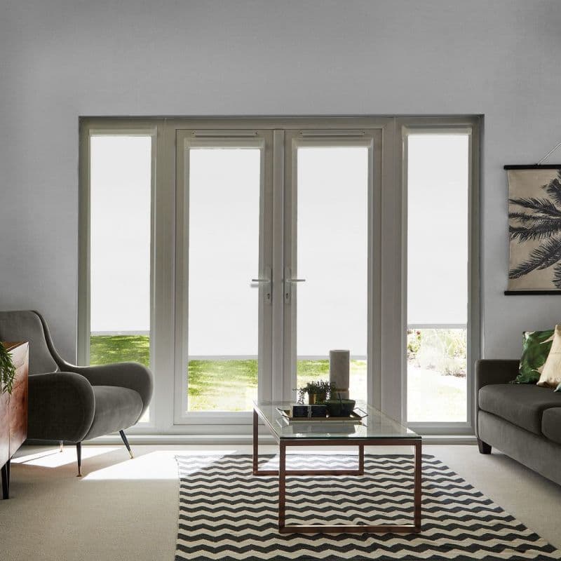 White roller blinds lowered over four tall glass doors and sidelights, filtering light with narrow bottom gaps; living room with grey seating, glass coffee table, chevron rug and lawn view.