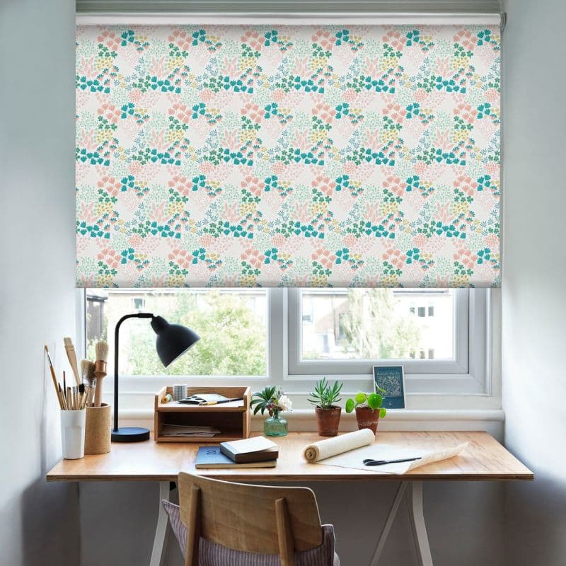 Floral roller blind (pastel pink, teal, green pattern), lowered and filtering daylight through the window; wooden desk beneath with lamp, potted plants, brushes and notebooks in a bright home study.