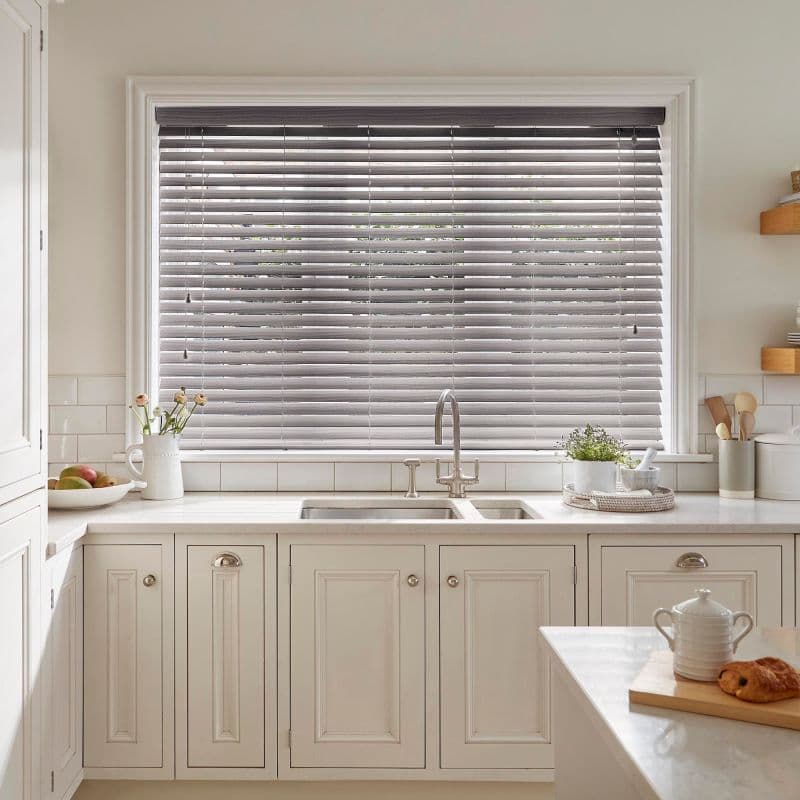 Gray horizontal venetian blinds lowered over a large window, slats partially tilted and filtering soft daylight; set above a cream kitchen sink with faucet, marble countertop, and small potted plants.
